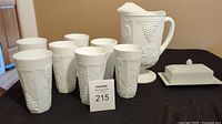 Set of milk glass pitcher, six tumblers, and ceramic butter dish arranged on table.