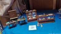 Overall lot showing two miniature wooden bakery display cases, two miniature black vintage-style sewing machines on wooden tables with spools of thread, and a small wooden fabric cabinet