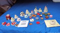 Image shows 23 miniature cakes and cake-themed miniature items arranged on a blue cloth background, including tiered wedding cakes, heart-shaped cakes, and assorted small cakes with floral and ribbon decorations.