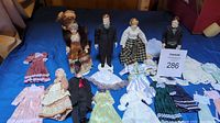 Seven assorted miniature Victorian dolls dressed in period clothing surrounded by various additional miniature dresses and garments laid out on a blue cloth.