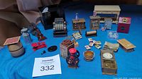 Full overview of all miniature general store accessories including register, scales, milk can, coffee grinder, furniture, and various small boxed items on blue cloth.