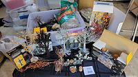 Overview of miniature Halloween and fall decorations including trees, small Halloween accessories, packaged Spanish moss, and bales of hay on a table with some items displayed inside boxes.