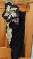 Full view of black off-shoulder dress hanging on door showing large gold and silver flower embellishment on shoulder and midsection draping.