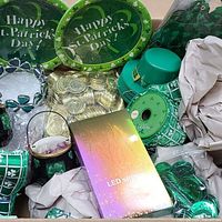 Box with green shamrock paper plates, green leprechaun hat, shamrock garland, gold foil coins, and sealed LED strip light package, surrounded by packing paper