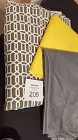 Three pieces of upholstery fabric including a grey geometric patterned piece, solid yellow piece, and solid grey piece, folded and stacked on a dark background.