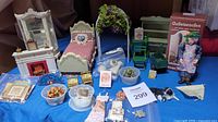 Overview of miniatures including Victorian bed, white fireplace, curio cabinet, garden arch, wood stove in box, gardener figurine, dog with puppies, and miniature accessories arranged on blue cloth