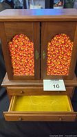 Front view of vintage wooden jewelry box with two doors featuring colorful floral panels, and a bottom drawer pulled open showing yellow lining. Wood finish shows light surface wear.