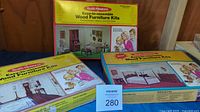 Three sealed Vintage Realife Miniatures Easy-to-Assemble Wood Furniture Kit boxes arranged side by side with different room themes: Entrance Hall, Living Room, and Bedroom. Boxes feature colorful illustrations and mid-century style family images.