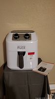 Photo of the white Wolfgang Puck air fryer with two control knobs and black handle, placed on a small table with a manual beside it.