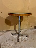 Side view of a round table showing the chrome base with four-pronged feet and central support column, wood table top edge visible beneath glass surface.