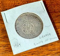 Top view of the 1964 Canadian silver dollar coin in a protective holder showing engravings and floral designs.