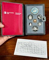 Open black textured box with red satin interior showing the full set of 7 coins in gray foam insert along with specification sheet on wooden surface