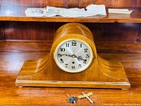 Side view of tambour mantel clock showing polished wooden case and round dial with hands, placed on wood surface with winding key and brass pendulum laid in front.