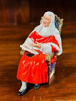 Side angle view of 'The Judge' Royal Doulton figurine in red robe seated on ornate chair with book in hand.
