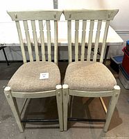 Front view of two antique white painted wooden counter height stools with beige fabric padded seats.