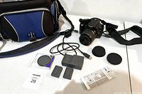 Complete camera kit with Canon Rebel XT camera fitted with 18-55mm lens, charger, two batteries, lens caps, USB cable, and blue and black carry bag.