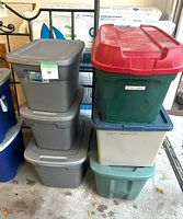 Stacked plastic storage bins: 3 gray bins on left, 3 bins on right including green Christmas bin with red lid, beige bin with wheels, and green bin with blue lid.