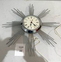 Front view of silver starburst wall clock showing round clock face with Roman numerals and thin rods radiating outward.
