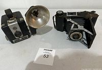 Two vintage cameras, one Kodak Brownie Flash, one folding bellows camera, shown side by side with flash attachment visible on Brownie Flash.