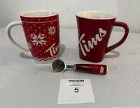 Front view of two ceramic Tim Hortons coffee mugs and a metal coffee scoop with a red handle showing Tim Hortons branding, arranged side by side on a white surface.