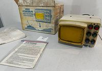 Vintage fish finder device with yellow screen and multiple control knobs, showing wear and discoloration, with the original box and instruction manual nearby.