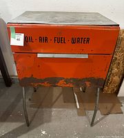 Front view of an orange vintage gas station service bin with text indicating oil, air, fuel, and water compartments, showing heavy wear and rust near the base.