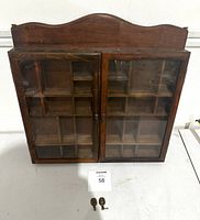 Front view of the wooden wall display cabinet showing glass paneled double doors and multiple small compartments inside. Two decorative metal wall hangers and key placed in front of the cabinet.