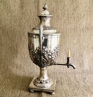 Front view of silver plated tea urn showing ornate repousse floral decoration, spout with wooden lever, and tall lid with finial.