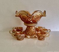 Front view of marigold carnival glass punch bowl on stand surrounded by six matching cups, showing iridescent finish and detailed embossing.