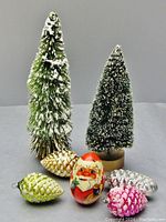 Two green bottle brush Christmas trees with white flocking snow effect; one tall at 9.5 inches and one smaller, along with a hand-painted Santa wooden egg and four pine cone-shaped glass ornaments in metallic colors with snow accents.