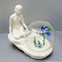 Front-left angle showing ceramic nude figure holding round glass fish bowl with art glass fish inside.