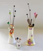 Photo of two hat pin holders and one stick pin holder displaying the pins from a front angle, showing floral decoration on holders and variety of pins.
