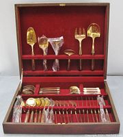 Opened wooden case showing top row of serving utensils including large spoons and cutters, all brass toned with wood handles.