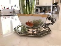 Side view of porcelain cup in silver holder on octagonal saucer showing fish motif and dragon handle