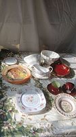 Photo showing assorted serving dishes including bowls, plates, sauce boat, and cups arranged on floral tablecloth with natural light
