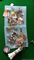 Top view of a collection of assorted cuff links, thimbles, keychains, pins, a shell brooch, a porcelain collectible figure, and a small metal ashtray arranged on blue fabric on a green surface.