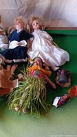Photo showing 5 dolls lying down including porcelain dolls in velvet and white dresses, and cloth doll in grass skirt with floral garlands