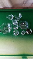 Overhead photo of ten assorted clear glass dishes and smallware on a green surface, showing different shapes and designs.