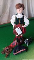 Porcelain doll in green velvet vest, white blouse and red tartan skirt on a wooden stand with black dog figurine and doll accessories on green surface
