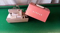 Photo showing the Singer Sew Handy mini electric sewing machine on its beige base with power cord and foot pedal, alongside the pink metal carrying case labeled 'SINGER'.