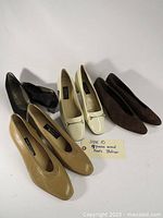 Four pairs of women's heels in beige leather, cream patent leather, brown suede, and dark brown/black leather or suede arranged together on white background. Includes handwritten note indicating size 10 and mixed leather materials.