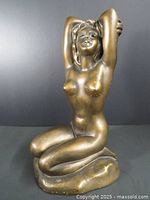 Front view of bronze-coloured kneeling nude woman sculpture showing the figure's face, arms raised behind head, and detailed torso.
