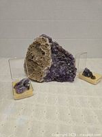 Wide shot showing the large amethyst geode with its natural rough edges and two smaller amethyst crystal specimens in plastic display cases with yellow foam.