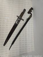 Two bayonets displayed side by side with scabbards on a textured white background.