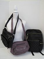 Photo showing three bags displayed on a white mannequin torso: a black faux leather handbag with multiple zippered compartments and a shoulder strap on the left, a mauve handbag with front pockets in the center, and a black quilted backpack on the right with an attached keychain.