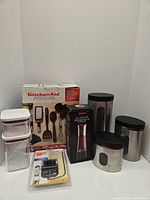 Overall view of the lot showing boxed KitchenAid kitchen gadget set, boxed Savora oil mister spray, OXO thermometer in packaging, three stainless steel storage containers with black lids and viewing windows, and two clear plastic rectangular food storage containers.
