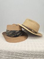 Photo showing two large, woven hats stacked together on a neutral background, highlighting their materials and bands.