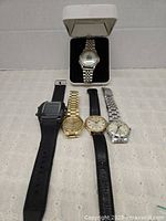 Five wristwatches arranged on a surface including one in original box with silver and gold-tone metal band, two with black leather straps, and two metal link band watches in gold and silver tones.