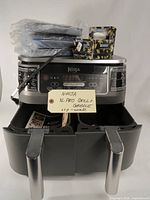 Front view of Ninja XL Pro Grill & Griddle appliance showing dual baskets pulled out with silver handles, control panel with digital buttons and display, black power cord, quick start guide, and accessories on top including a small boxed item and plastic wrapped parts.