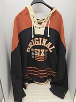Front view of the black and burnt orange lace-up hockey hoodie showing the Original Six text and NHL team logos embroidered on chest.